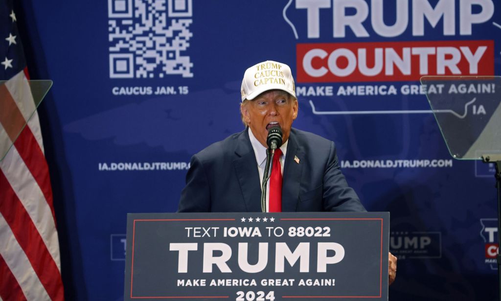 Donald Trump wears a suit and tie with a hat that reads 'Trump caucus captain' as he speaks to a crowd at the 2024 Iowa caucus