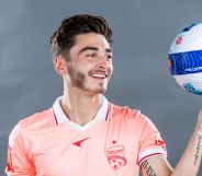 Josh Cavallo wears a pale pink Adelaide United football uniform as he smiles and holds up a blue, red and white football in one hand