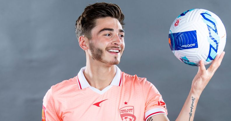 Josh Cavallo wears a pale pink Adelaide United football uniform as he smiles and holds up a blue, red and white football in one hand
