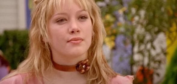 A still of Hilary Duff wearing a pink shirt and copper colour choker with a flower on it as she portrays the Disney Channel character Lizzie McGuire