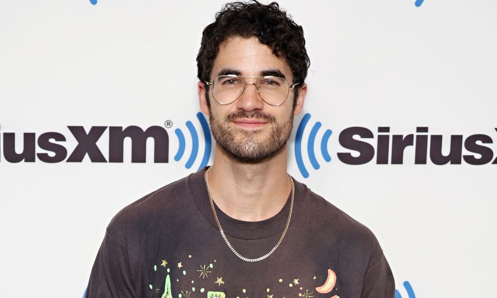 Darren Criss, a non-LGBTQ+ actor who got famous for playing queer roles, wears a brown shirt, necklace and glasses as he smiles for the camera