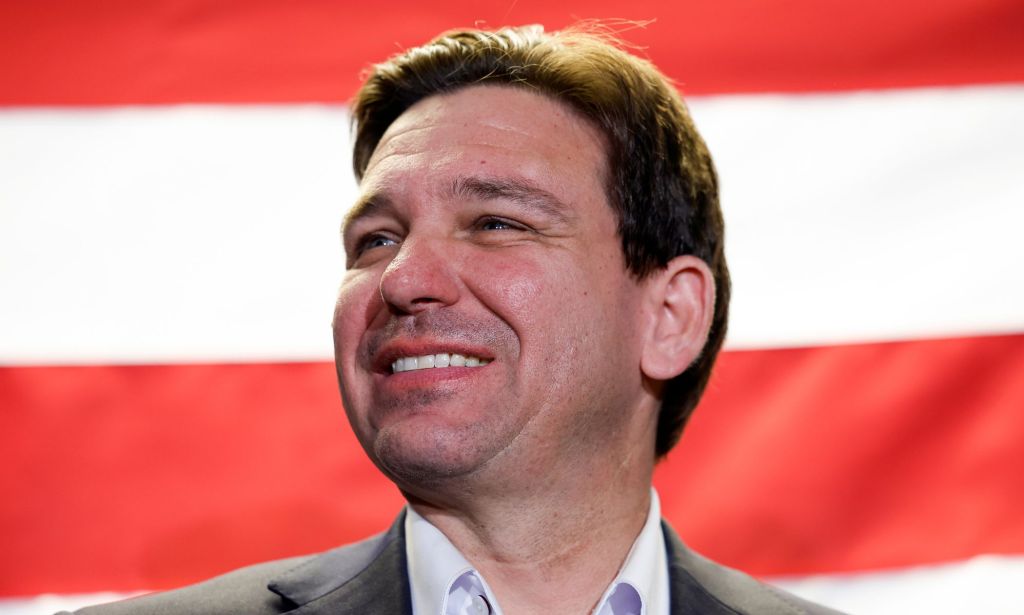 Florida governor Ron DeSantis, who once was a top candidate in the 2024 Republican presidential race, smiles awkwardly as he stands in front of the red and white stripes of the US flag