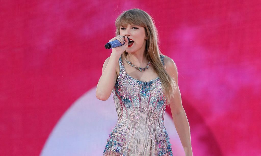 Taylor Swift wears a silver sparkly outfit while she sings into a microphone during a concert. Her team is reportedly furious after the New York Times published an op-ed that claimed Taylor Swift had been hinting she's queer throughout her career