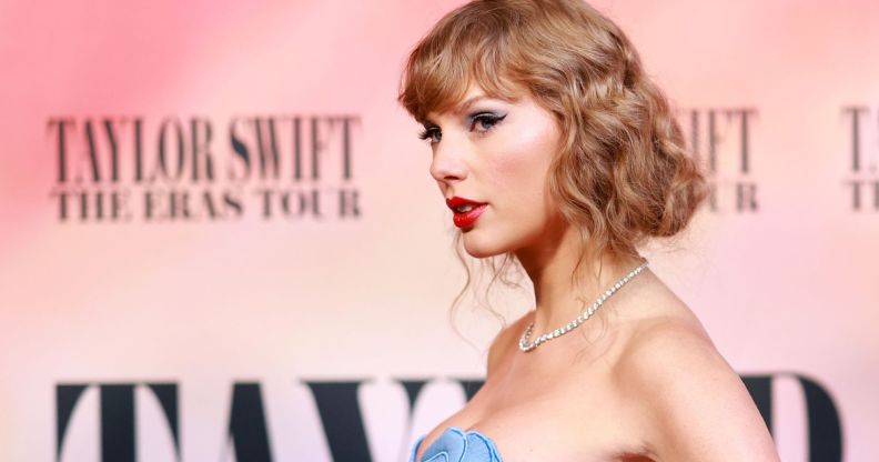 Taylor Swift wears a blue dress while standing in front of a background for her Eras tour. Her team is reportedly furious after the New York Times published an op-ed that claimed Taylor Swift had been hinting she's queer throughout her career