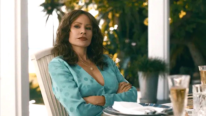 A photo of Sofia Vegara playing Griselda Blanco, she is sitting at a table outdoors with her arms folded and wearing a blue dress. She has long dark hair.