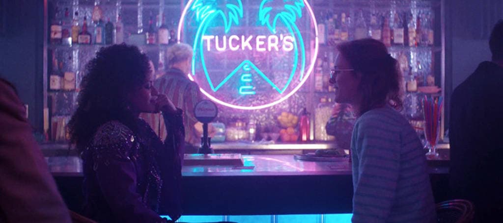Scene from the Black Mirror episode 'San Junipero' featuring two women sitting at a bar, the lighting is pink and blue