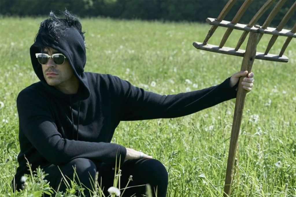 David Rose hides in a field with a pitchfork he's wearing sunglasses and a black hoody