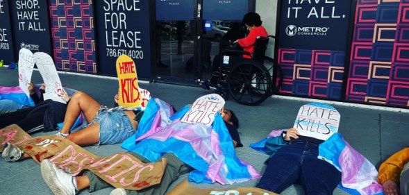 Image shows people lying on a pavement draped in trans flags and holding up cardboard tombstones that read 'hate kills'