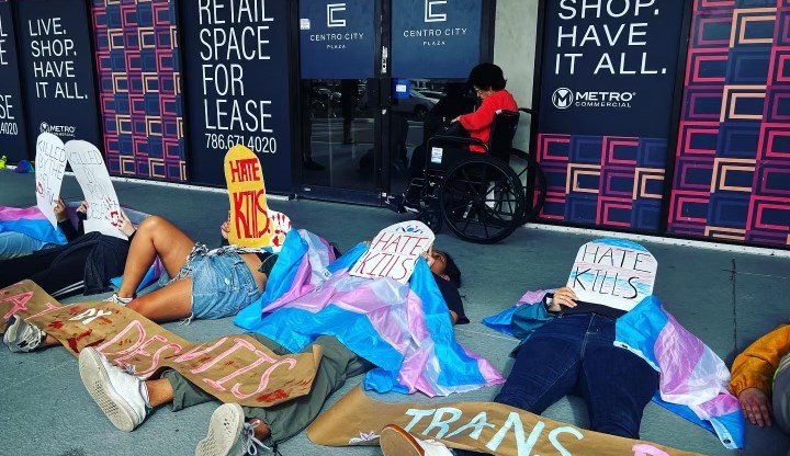 Image shows people lying on a pavement draped in trans flags and holding up cardboard tombstones that read 'hate kills'