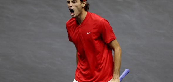 Tennis player Taylor Fritz shouts while holding his racquet on court, he is wearing a red tee shirt and matching headband.