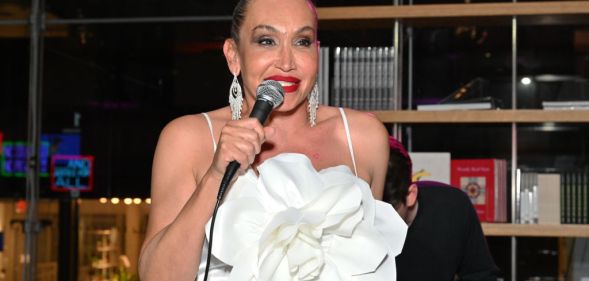 Image shows trans activist Cecilia Gentili at an event, wearing a white dress and holding a microphone