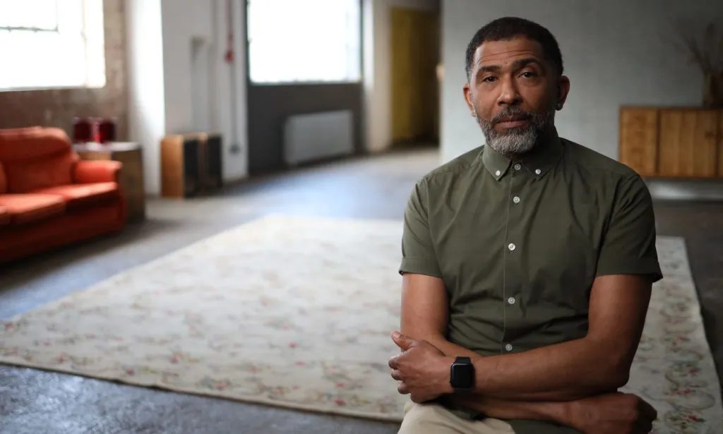 This is an image of a Black man in an interview setting. He is wearing a short sleeve green shirt and his arms are folded.