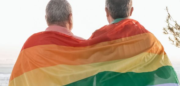 Two elderly people hudle together under a Pride flag.