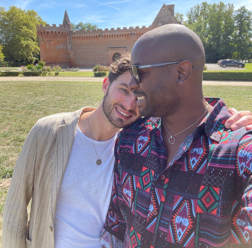 This is an image of two men embracing each other. The man on the left is white and is wearing a beige cardigan and white t-shirt. The man on the right is Black and is wearing an aztec print shirt and dark sunglasses. 
