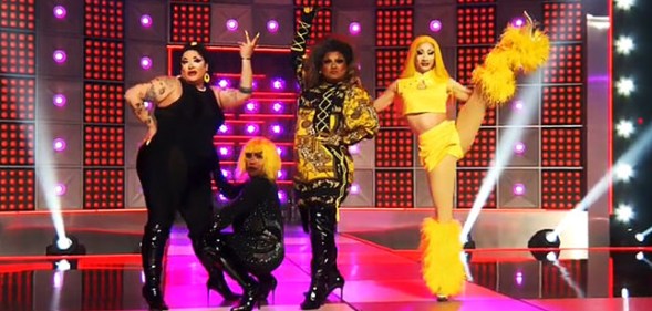 Image shows four Drag Race queens on stage doing the girl group challenge, wearing flamboyant costumes. One is high kicking