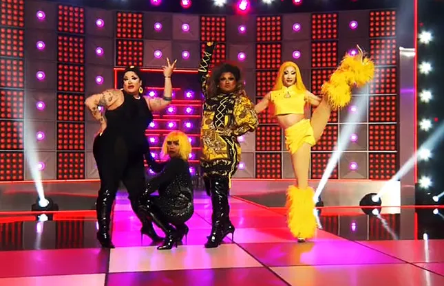 Image shows four Drag Race queens on stage doing the girl group challenge, wearing flamboyant costumes. One is high kicking