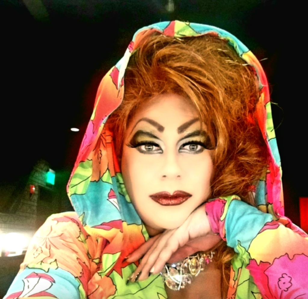 Image shows Nashville drag queen Veronika Electronika in a colourful outfit with a hood pulled up over her hair, she is resting her head on her hand and looking into the camera. 