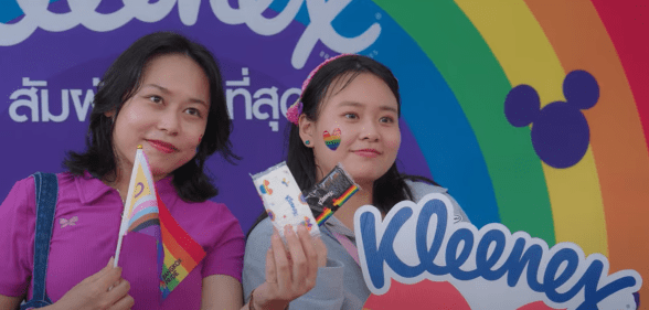 Image shows two people posing at a Thai Pride event in front of the Kleenex brand logo, both have rainbow stickers on their cheeks.