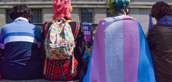 A group of people sit with their backs to the camera, one wearing a trans flag as a cape.