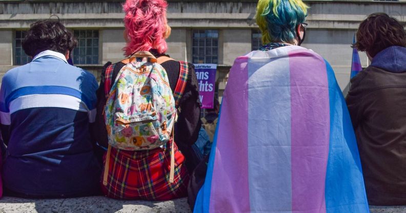 A group of people sit with their backs to the camera, one wearing a trans flag as a cape.