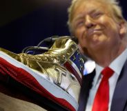 Donald Trump wears a suit and tie while smiling and standing in front of a gold-coloured high top sneaker with a T emblazoned on it and a US flag print