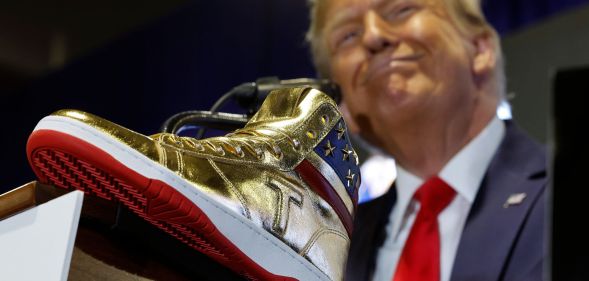 Donald Trump wears a suit and tie while smiling and standing in front of a gold-coloured high top sneaker with a T emblazoned on it and a US flag print
