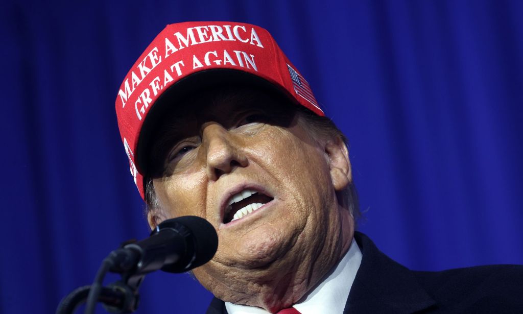 Former president Donald Trump wears a suit and tie and a red "Make America Great Again" hat while speaking into a microphone