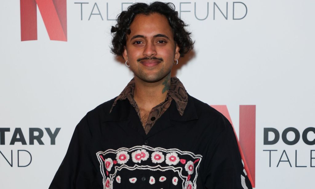 Krishna Istha, a comedian featured on Hannah Gadsby's Netflix special Gender Agenda, wears a dark shirt as they stand in front of a background with the Netflix logo on it