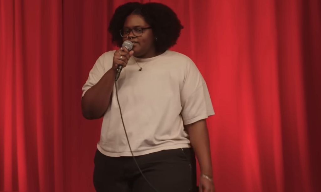Asha Ward, a comedian featured on Hannah Gadsby's Netflix special Gender Agenda, wears a light shirt and dark trousers while speaking into a microphone during a performance