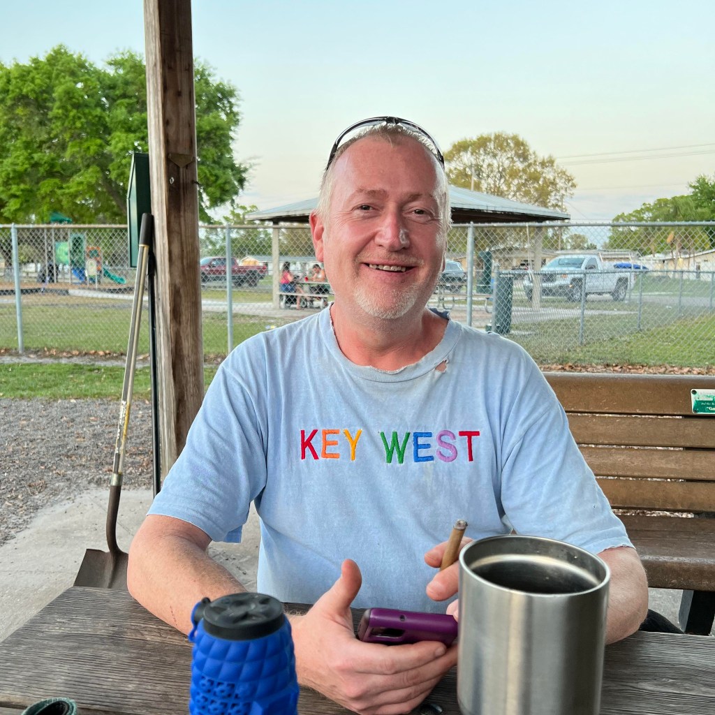 John Walter Lay smiling in the dog park wearing a Key West T-shirt, each letter is in a different colour of the rainbow. 