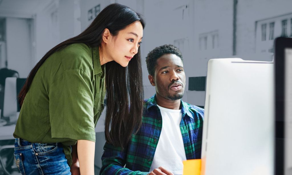 This is an image of an Asian woman and a Black man. She is wearing green and is standing while the man is sitting. They are looking at a computer screen.