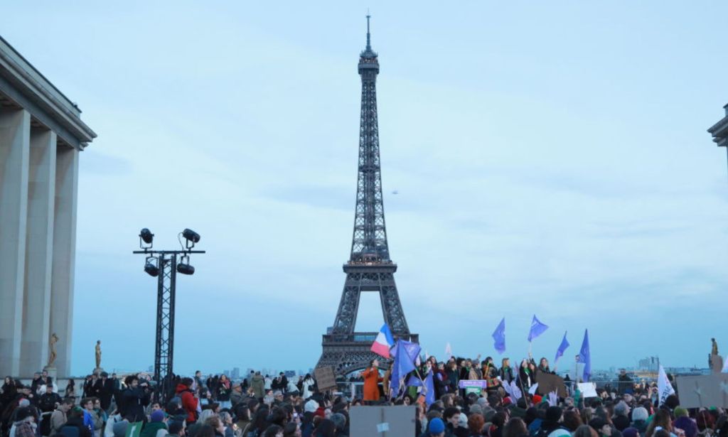 This is an image of the Eifel Tower in Paris, France. It is an overcast day. There is a large group of people in the foreground and we can see French flags flying.