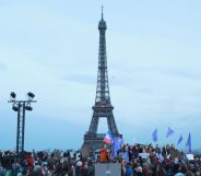 This is an image of the Eifel Tower in Paris, France. It is an overcast day. There is a large group of people in the foreground and we can see French flags flying.