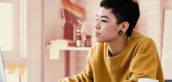 This is an image of a person working at a computer. They present genderqueer. They have large gauge earrings and are wearing a yellow jumper.