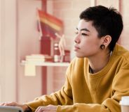 This is an image of a person working at a computer. They present genderqueer. They have large gauge earrings and are wearing a yellow jumper.