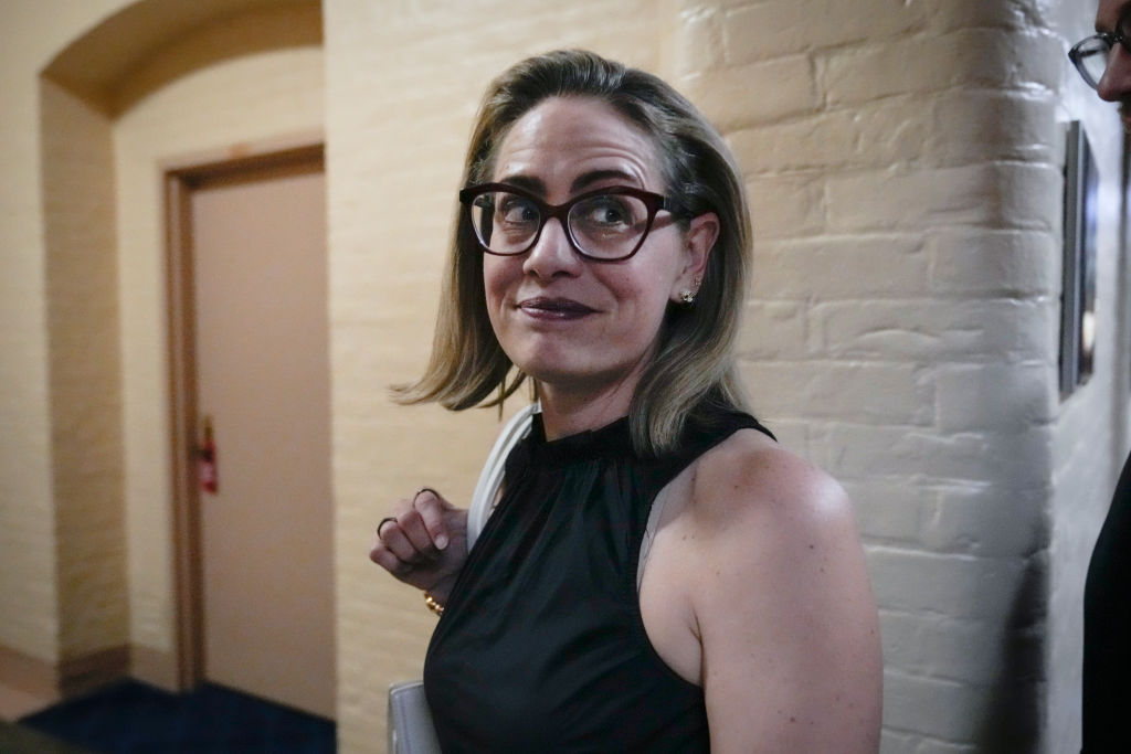 Kyrsten Sinema in a white brick hallway, turning towards the camera with a white bag over her far shoulder. She's wearing glasses and a black sleeveless top.