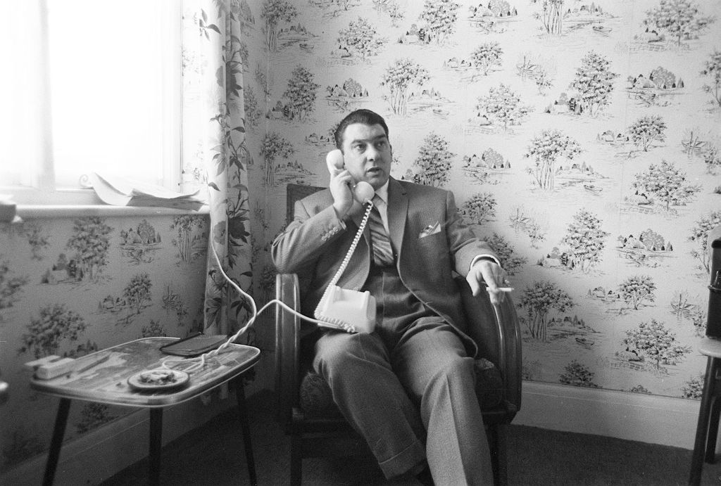 Ronnie Kray sits at home, smoking and holding a phone to his ear.
