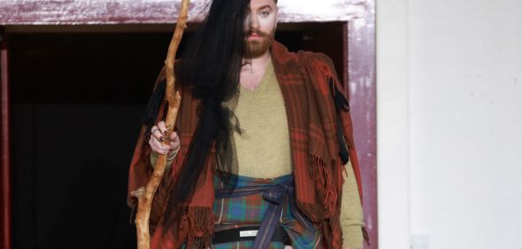 Non-binary singer Sam smith pictured at Paris Fashion Week wearing a tartan shall, a mini kilt, with long hair extensions and carrying a wooden staff