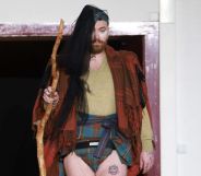 Non-binary singer Sam smith pictured at Paris Fashion Week wearing a tartan shall, a mini kilt, with long hair extensions and carrying a wooden staff