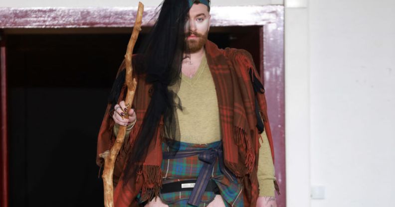Non-binary singer Sam smith pictured at Paris Fashion Week wearing a tartan shall, a mini kilt, with long hair extensions and carrying a wooden staff