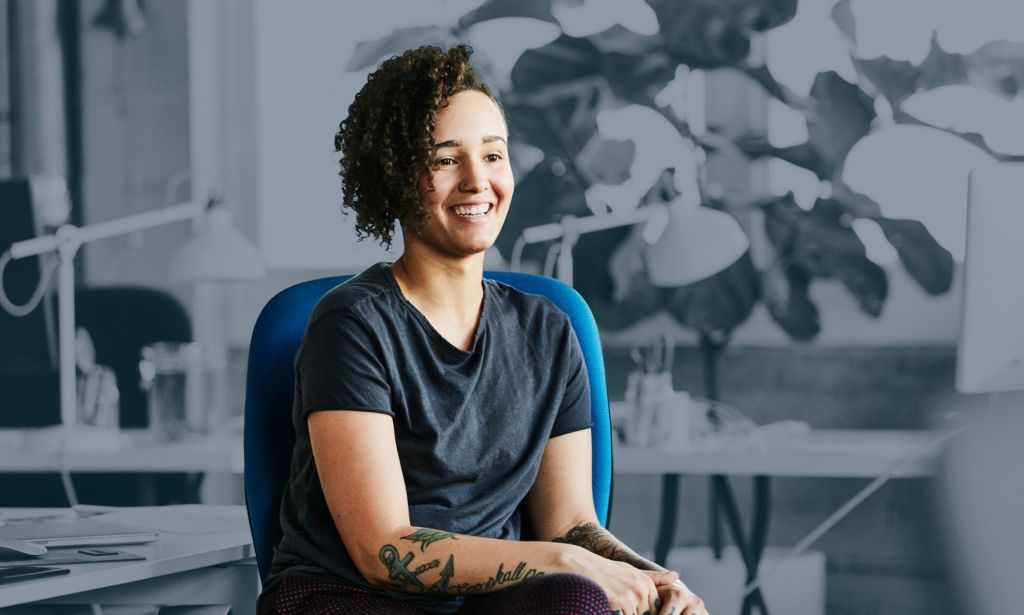 This is an image of a female presenting person sitting on a chair. They are wearing a dark tshirt and have various visible tattoos. She is smiling. She is in full colour while the rest of the image is in black and white. 