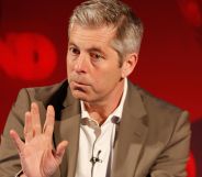 Justin Webb waving his hand during a speech on stage at a press event.