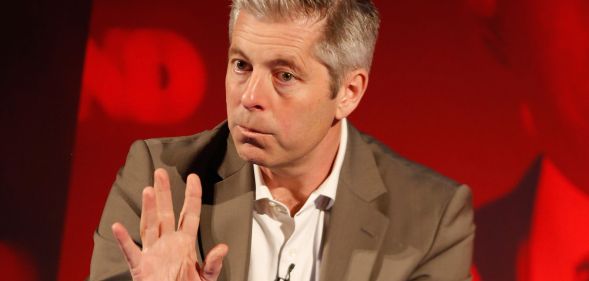 Justin Webb waving his hand during a speech on stage at a press event.
