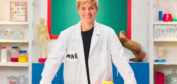 Mae Martin on the set of Fluid: Beyond the Binary, wearing a labocoat and smiling.