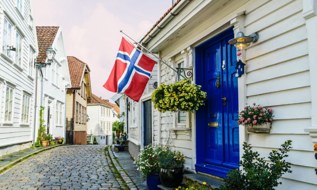 This is an image of the Norwegian flag on a street. The flag is red with a blue cross with white outlines and it is hung outside a blue door on a small village street.