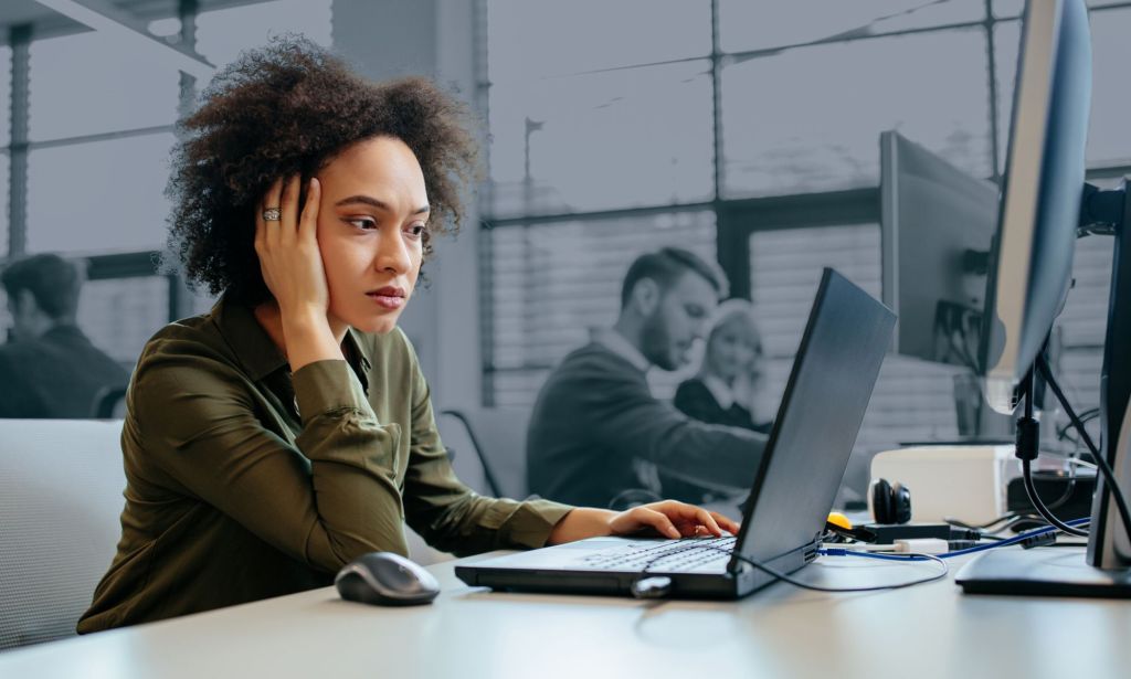 This is an image of a Black woman looking stressed at her work computer. She is in full colour while the rest of the image is in black and white. 