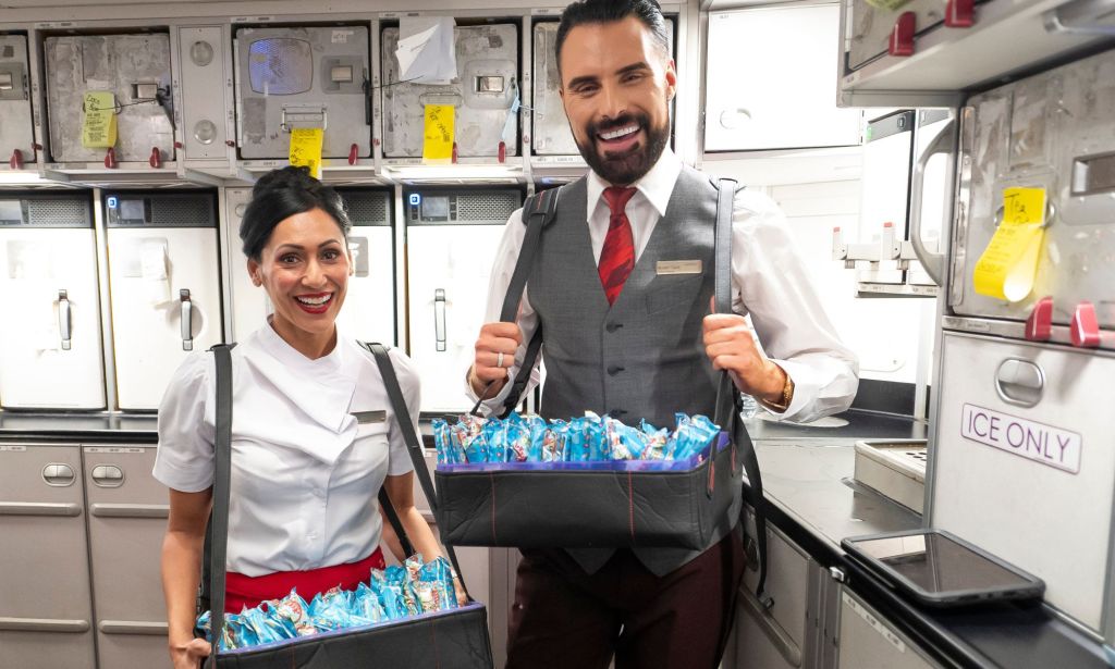 This is an image of celebrity Rylan Clark standing next to a woman in the galley of an airplane. They are wearing Virgin Atlantic's cabin crew uniform and have trays of snacks.