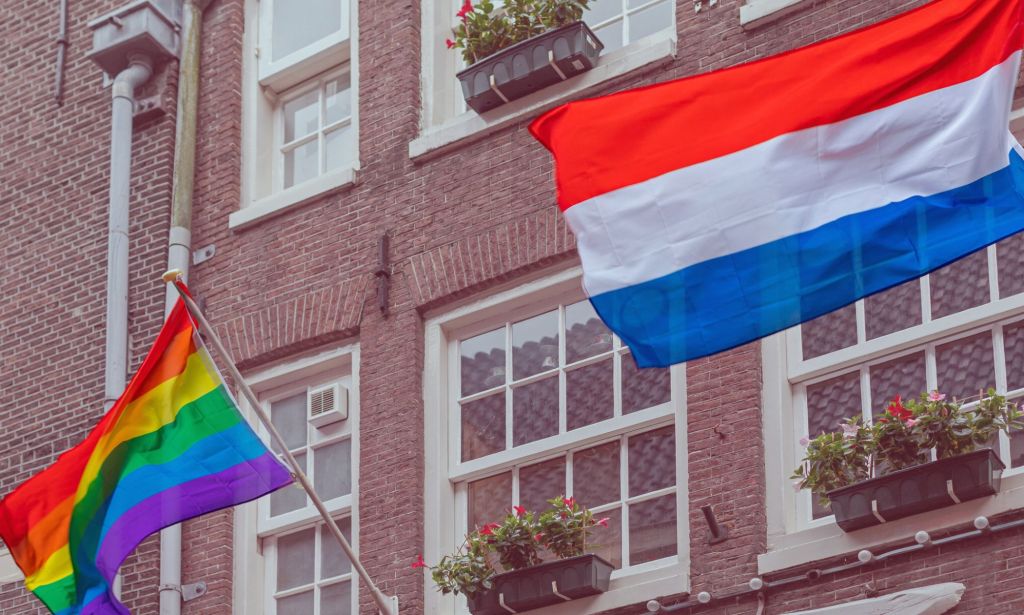 This is an image of the Pride flag flying next to the Netherlands flag on a building in Amsterdam. 