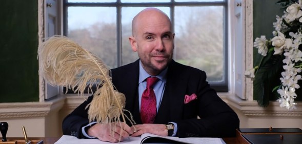 Tom Allen sat at a wooden desk, a window behind him, signing wedding documents with a very large quill.