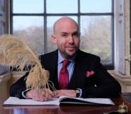 Tom Allen sat at a wooden desk, a window behind him, signing wedding documents with a very large quill.